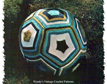 Puede incluir: Una pelota de crochet con un patrón geométrico en tonos de azul, verde, blanco y negro. La pelota está sentada sobre un lecho de hierba verde.