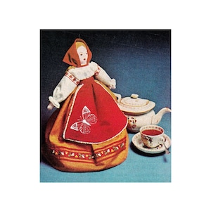 Può includere: Un copri-teiera rosso e dorato a forma di bambola che indossa un vestito rosso e un grembiule bianco con una farfalla ricamata sul davanti. La bambola ha un cappuccio rosso e un viso bianco.