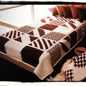 May include: A handmade, geometric-patterned blanket in shades of brown and cream, displayed on a bed. The blanket features a patchwork design with triangles, squares, and striped patterns. The bed is adorned with brown pillows, and a matching throw is draped over the side.