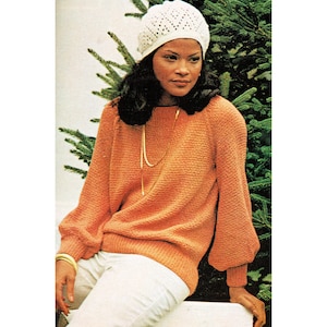 Könnte beinhalten: Eine Frau trägt einen weißen Häkelhut und einen leuchtend orangenen Pullover mit langen Ärmeln. Sie sitzt auf einer weißen Oberfläche mit einem grünen Baum hinter ihr.