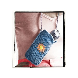 Peut inclure: Un étui à lunettes en tricot bleu avec un motif de fleur brodé jaune et orange. L'étui est suspendu au cou d'une personne par un cordon violet et blanc.