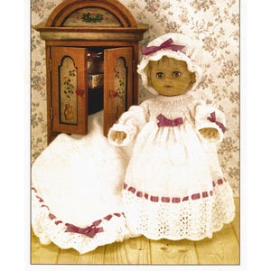 Puede incluir: Un vestido de muñeca de punto blanco con una cinta rosa y un gorro de punto blanco a juego. La muñeca está sentada frente a un armario de madera con un fondo de papel tapiz floral. El texto "It's bedtime" está escrito debajo de la imagen.