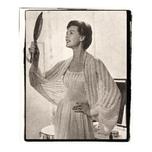 May include: A black and white photo of a woman wearing a white dress and a crocheted shawl. She is holding a hand mirror and looking at her reflection.
