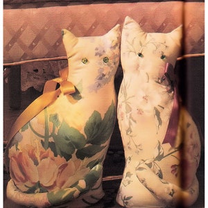 Puede incluir: Dos juguetes de peluche con forma de gato con patrones florales. Un gato es marrón claro con hojas verdes y una cinta amarilla. El otro gato es blanco con flores rosas y una cinta rosa.