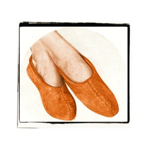 以下が含まれることがあります： ステッチのディテールが施されたオレンジ色のスエードスリッパのペア。スリッパは足のペアに履かれています。