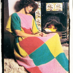May include: A woman wearing a colorful patchwork dress with a pink, yellow, green, and red pattern. She is sitting in front of a fireplace with a black tiled surround.