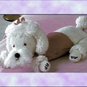 May include: A white knitted poodle toy with a pink bow and a pink and white striped body. The toy is lying on a pink blanket.