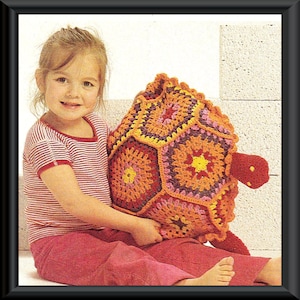 Peut inclure: Une jeune fille tient un coussin en forme de tortue crocheté. Le coussin est fait de fil coloré dans des tons d'orange, jaune, rose et violet. La tortue a une carapace faite de six formes hexagonales.