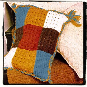 May include: A crocheted pillow with a patchwork design in shades of blue, brown, orange, and white. The pillow has a crocheted border in a contrasting color.