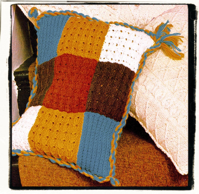 May include: A crocheted pillow with a patchwork design in shades of blue, brown, orange, and white. The pillow has a crocheted border in a contrasting color.