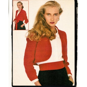 May include: A red cropped jacket with a white blouse and a black skirt. The jacket has a tie at the waist and the model is wearing a gold bracelet.