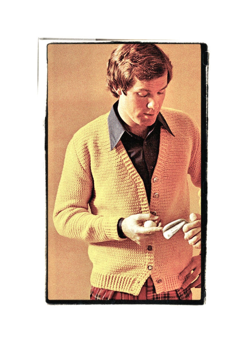 May include: A man wearing a yellow knitted cardigan with a button-up collar and a plaid shirt underneath. He is holding a golf ball and a golf club.