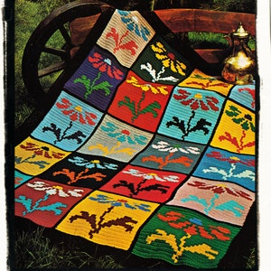 Puede incluir: Una manta de ganchillo colorida con un patrón floral. La manta está hecha de cuadrados con flores y hojas de diferentes colores. La manta está drapeada sobre un banco de madera con una jarra de metal al lado.