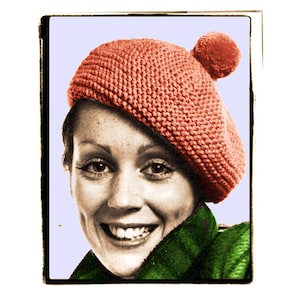 May include: A woman wearing a red knitted beret with a pom-pom on top. She is smiling and wearing a green scarf.