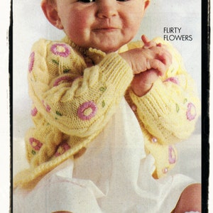 May include: A baby wearing a yellow knitted cardigan with pink flower embroidery. The cardigan has a button closure and is worn over a white dress. The baby is sitting on a white surface. The text "FLIRTY FLOWERS" is printed in the top right corner of the image.