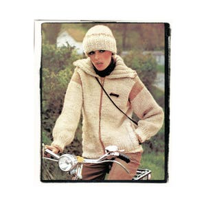 May include: A woman wearing a cream-colored knitted jacket with brown accents, a cream-colored knitted hat, and brown pants. She is riding a bicycle.