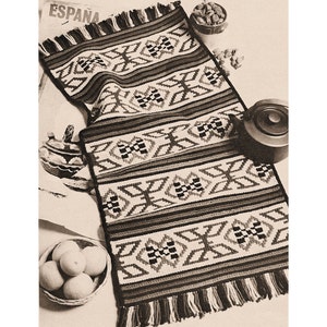Puede incluir: Una alfombra de crochet negra, blanca y marrón con un patrón geométrico. La alfombra tiene flecos en los extremos y está sobre una mesa con fruta y otros objetos.