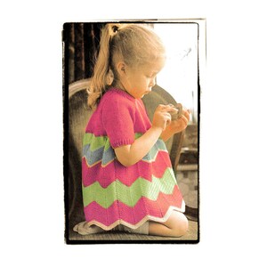 May include: A young girl wearing a pink, green, and blue striped knitted dress. The dress has a chevron pattern and is a little longer in the back than the front.