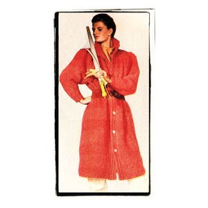 May include: A red knitted cardigan with a long, button-up front and large, puffed sleeves. The cardigan is belted at the waist and the model is holding a long, thin object in her hand.