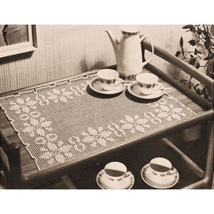 May include: A black and white photo of a crocheted table runner with a floral design. The runner is on a wooden table with a teapot and four cups and saucers.