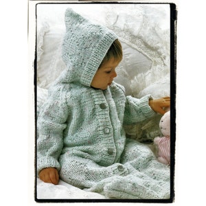 May include: A baby wearing a light green knitted hooded jumpsuit with white buttons. The jumpsuit has a cable knit pattern and is decorated with small white dots. The baby is sitting on a white bed and holding a pink and white striped stuffed animal.