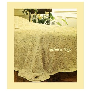 May include: A cream-colored bedspread with a detailed, textured pattern covers a bed. The design features repeating fan-like shapes and geometric patterns. The text "Yesterdays Magic" is visible on the bedspread. The bedspread is on a wooden floor.