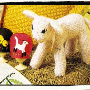 May include: A knitted white lamb toy with black eyes. The lamb is sitting on a bed of straw with two small knitted hats in the foreground.