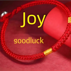 以下が含まれることがあります： 赤い編み込みブレスレット、銀色のベルチャームと金のアクセント付き。「Joy」の文字が黄色で書かれ、下に「goodluck」と書かれています。ブレスレットは赤い表面にあります。