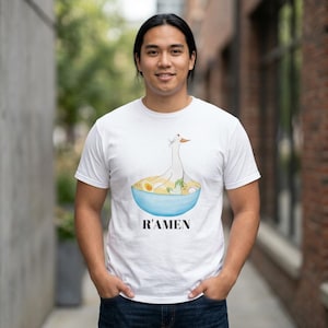 May include: A white short-sleeved t-shirt featuring a graphic of a white bird sitting in a blue bowl of ramen noodles with an egg and greens. The text R'AMEN is printed in black capital letters directly below the blue bowl illustration.