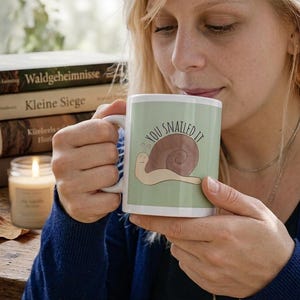 Könnte beinhalten: Eine weiße Keramiktasse mit hellgrünem Hintergrund und dem Aufdruck "YOU SNAILED IT". Auf der Tasse befindet sich auch eine Cartoon-Schnecken-Illustration. Die Tasse wird von einer Person gehalten.