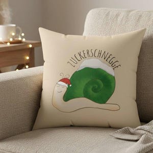 Könnte beinhalten: Beiges quadratisches Kissen mit einer Cartoon-Schnecke mit grünem Gehäuse und Weihnachtsmütze. Die Schnecke hat ein lächelndes Gesicht und den Text "Zuckerschnegge" darüber. Das Kissen liegt auf einem beigen Sofa.
