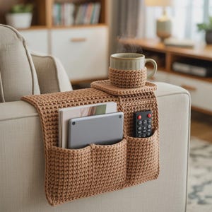 May include: A crocheted sofa armrest organizer in a light brown color. It features pockets for a remote, tablet, and books, with a built-in coaster and a mug. The mug has a matching cozy. The armrest is placed on a beige sofa.