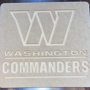May include: A light brown wooden sign featuring the Washington Commanders logo and team name. The logo is a large, stylized "W" and the words "WASHINGTON COMMANDERS" are carved into the wood. The sign is rectangular with rounded corners.
