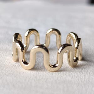 Può includere: Un anello color oro caratterizzato da un motivo ondulato o serpentino continuo e ripetitivo. La fascia è realizzata in un unico pezzo di metallo lucido formato da una serie di archi lisci e arrotondati che creano un design aperto.