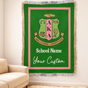 Op de afbeelding: Groene plaid met een roze rand en franjes. Het midden heeft een wapen met de letters AKA, een schoolnaam en de woorden "Your Custom" in een scriptlettertype. De deken wordt aan een muur getoond.