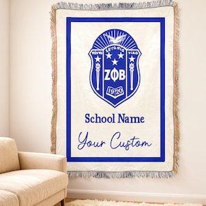Op de afbeelding: Een witte geweven sprei met een blauwe rand en een blauw wapenontwerp. Het wapen bevat tekst en de woorden "School Name" en "Your Custom" in blauw script. De deken heeft een gefranjerde rand.