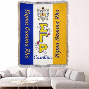 Op de afbeelding: Een geweven sprei met blauwe, witte en gouden verticale strepen. De sprei heeft de Griekse letters ΣΓΡ in goud, de woorden "Sigma Gamma Rho" in blauw en de naam "Caroline" in blauw. De sprei heeft een gefranjerde rand.