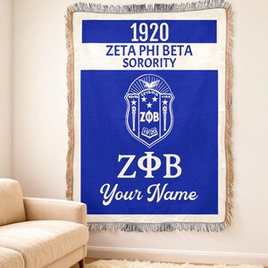 Op de afbeelding: Een blauw-witte Zeta Phi Beta Sorority-worpdeken. De deken heeft het wapen van de vereniging, het jaar 1920 en de tekst "Your Name". De deken heeft een gefranjerde rand en wordt aan een muur getoond.