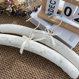 May include: Two padded clothing hangers covered in white fabric. The front hanger features delicate beige floral embroidery and a small cream-colored ribbon bow at the base of the metal hook. The back hanger is plain white fabric with a matching ribbon bow.
