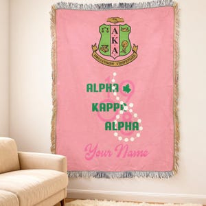 Op de afbeelding: Roze sprei met een gefranjerde rand. De deken heeft een wapen met groene en gouden accenten, de Griekse letters Alpha Kappa Alpha en het jaar 1908. De woorden "Your Name" zijn in roze gedrukt.