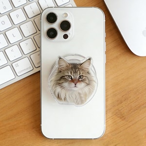 May include: A white smartphone with a clear phone grip featuring a detailed illustration of a fluffy cat's face. The cat has brown and gray fur, with a focused gaze. A keyboard and a computer mouse are in the background.