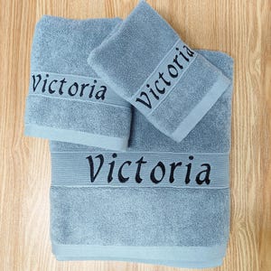 May include: A set of three pale blue bath towels, each embroidered with the name "Victoria" in black. The towels are arranged on a wooden surface. The set includes a large bath towel, a hand towel, and a face cloth.