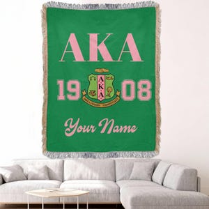 Op de afbeelding: Groene geweven sprei met roze tekst en een wapen. De sprei heeft de letters "AKA" boven de cijfers "1908" en de woorden "Your Name". De sprei heeft een gefranjerde rand.