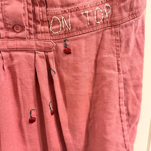 Puede incluir: Primer plano de una prenda de color rosa polvoriento con el texto bordado "ON TOP". La prenda presenta peque&ntilde;os dise&ntilde;os de cerezas bordadas y detalles de botones. La tela tiene una apariencia texturizada.