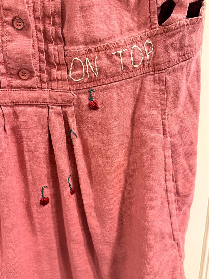 Puede incluir: Primer plano de una prenda de color rosa polvoriento con el texto bordado "ON TOP". La prenda presenta peque&ntilde;os dise&ntilde;os de cerezas bordadas y detalles de botones. La tela tiene una apariencia texturizada.