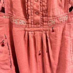 Puede incluir: Un mono de color rosa polvoriento con la frase bordada "THE CHERRY ON TOP". El mono presenta una parte delantera abotonada, pliegues y peque&ntilde;as cerezas bordadas. El mono tiene un bolsillo en el lado izquierdo.