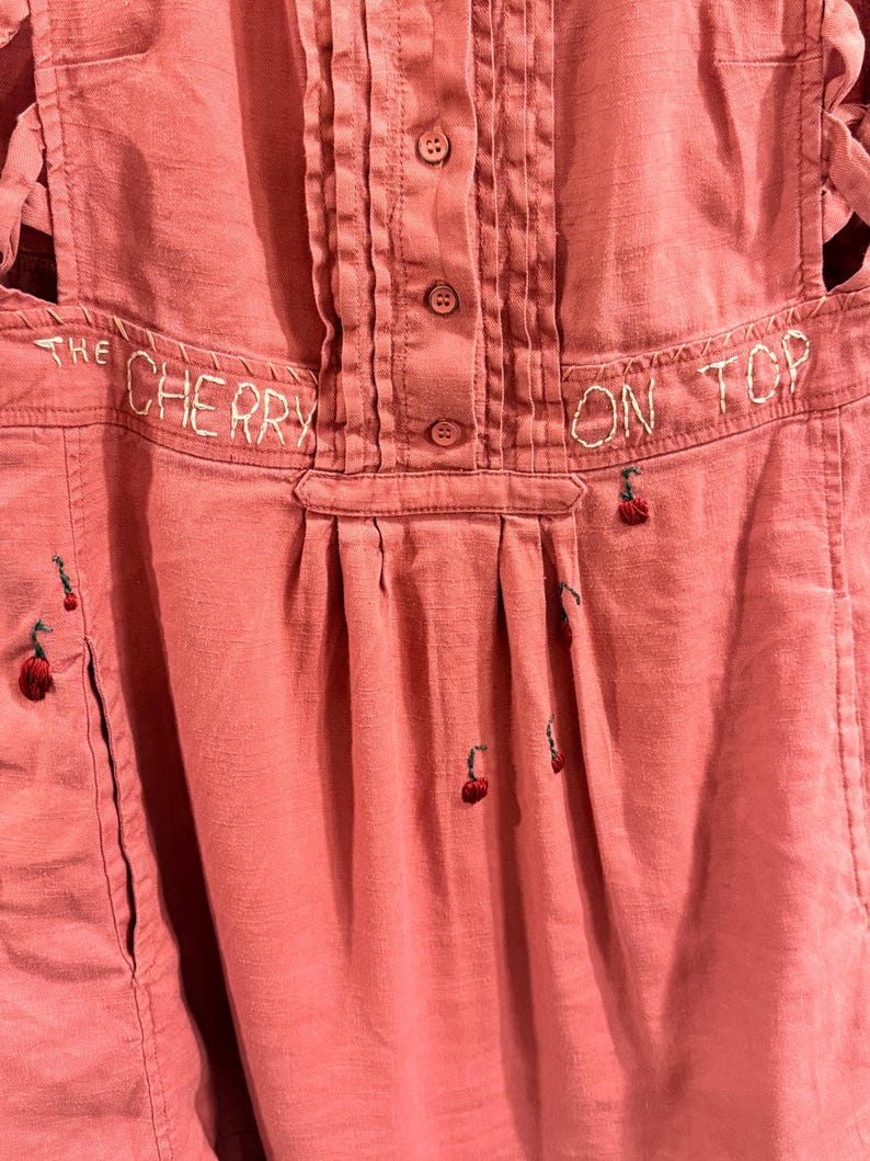 Puede incluir: Un mono de color rosa polvoriento con la frase bordada "THE CHERRY ON TOP". El mono presenta una parte delantera abotonada, pliegues y peque&ntilde;as cerezas bordadas. El mono tiene un bolsillo en el lado izquierdo.