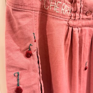 Puede incluir: Primer plano de una prenda de color rosa polvoriento con el texto bordado "THE CHERRY". La prenda presenta dise&ntilde;os de cerezas cosidos a mano con tallos verdes y cerezas rojas. La tela tiene un aspecto texturizado.