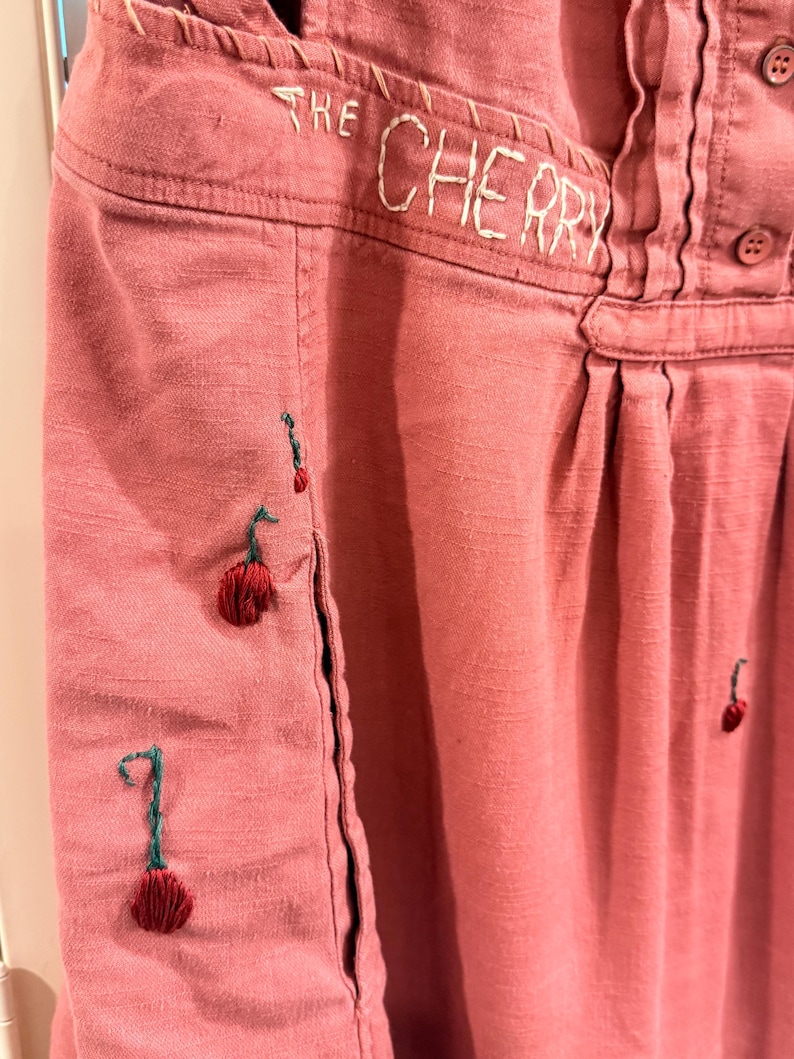 Puede incluir: Primer plano de una prenda de color rosa polvoriento con el texto bordado "THE CHERRY". La prenda presenta dise&ntilde;os de cerezas cosidos a mano con tallos verdes y cerezas rojas. La tela tiene un aspecto texturizado.