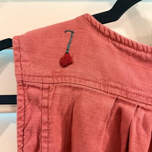Puede incluir: Primer plano de una prenda de color rosa empolvado con un peque&ntilde;o dise&ntilde;o floral bordado en rojo y verde en el hombro. La tela tiene una textura visible y est&aacute; colgada de una percha negra.